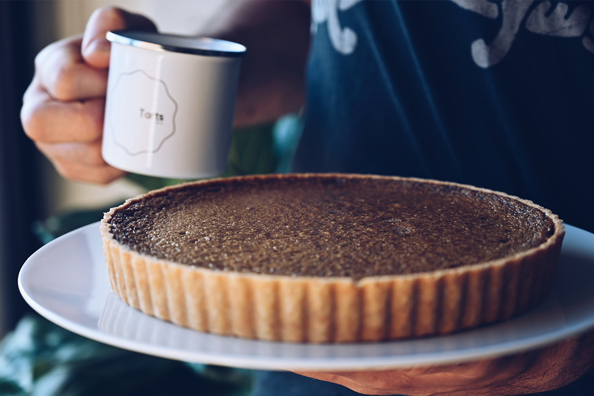 Baker holds coffee mug while carrying delicious rustic-looking tart on plate (tarts anon)