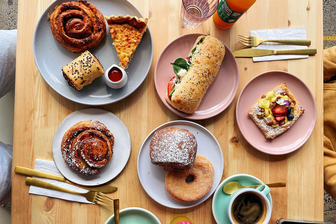 a wooden table with a brunch spread on different pastel plates, there are various sweet and savoury baked goods and a cup of coffee as well as cutlery (miss sina)