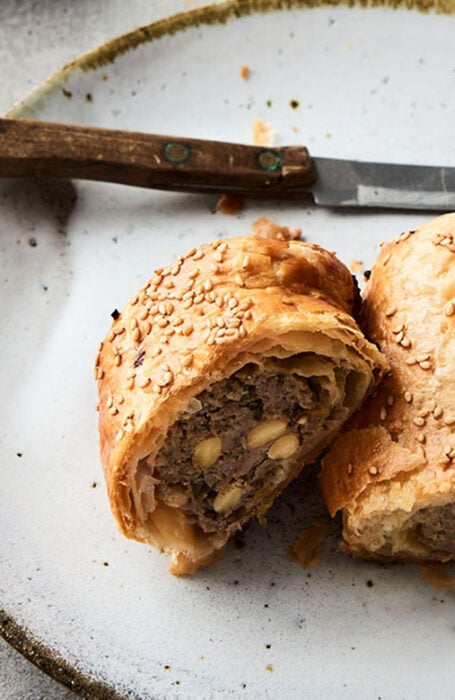 Delicious-looking sausage roll that has been cut in half width-ways sits on a plate next to a knife (pork, sage and onion sausage roll)