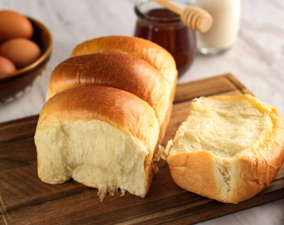 a fresh golden loaf of milk bread rests on a wooden bread board, it looks golden on the crust and soft and fluffy in the middle. one portion has been torn off and set beside the loaf on the board. in the background are a bowl of eggs, a jar of honey with a honey drizzler and a milk bottle (shokupan)