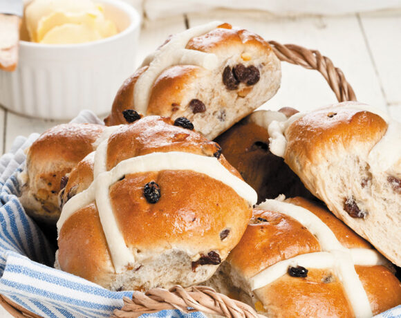 A basket full of hot cross buns sits on a counter in front of a bowl full of butter (bun spice)