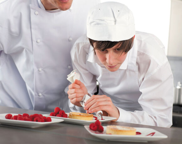 A pastry chef apprentice stands piping a coulis onto a plate which holds a tart and a pile of raspberries (apprenticeship)