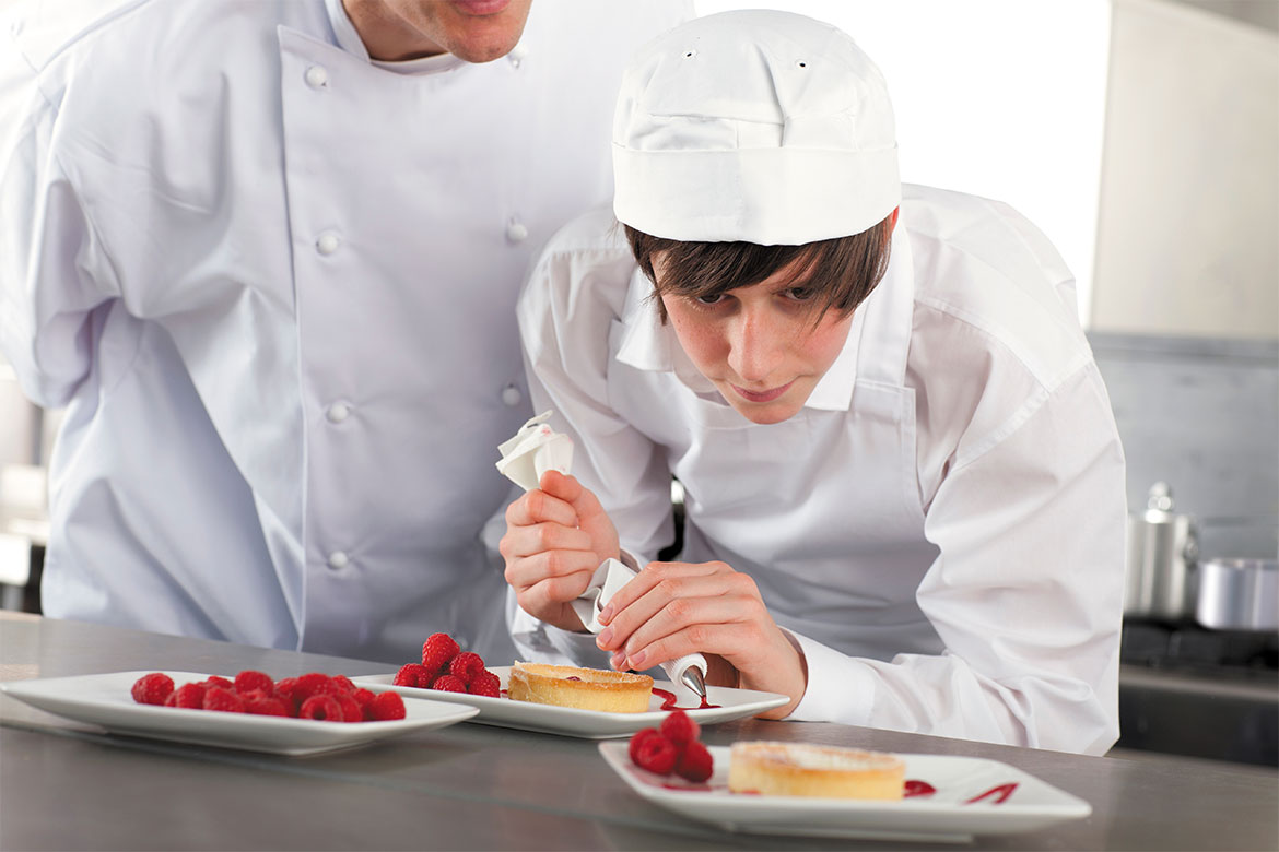 A pastry chef apprentice stands piping a coulis onto a plate which holds a tart and a pile of raspberries (apprenticeship)