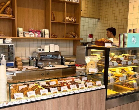 The interior of a bakery/cafe; there are big glass cases full of sweet and savoury pastries. Behind is a shelf stacked with bread and a coffee machine. On the right, behind the counter, there is a man. (Penny for Pound)