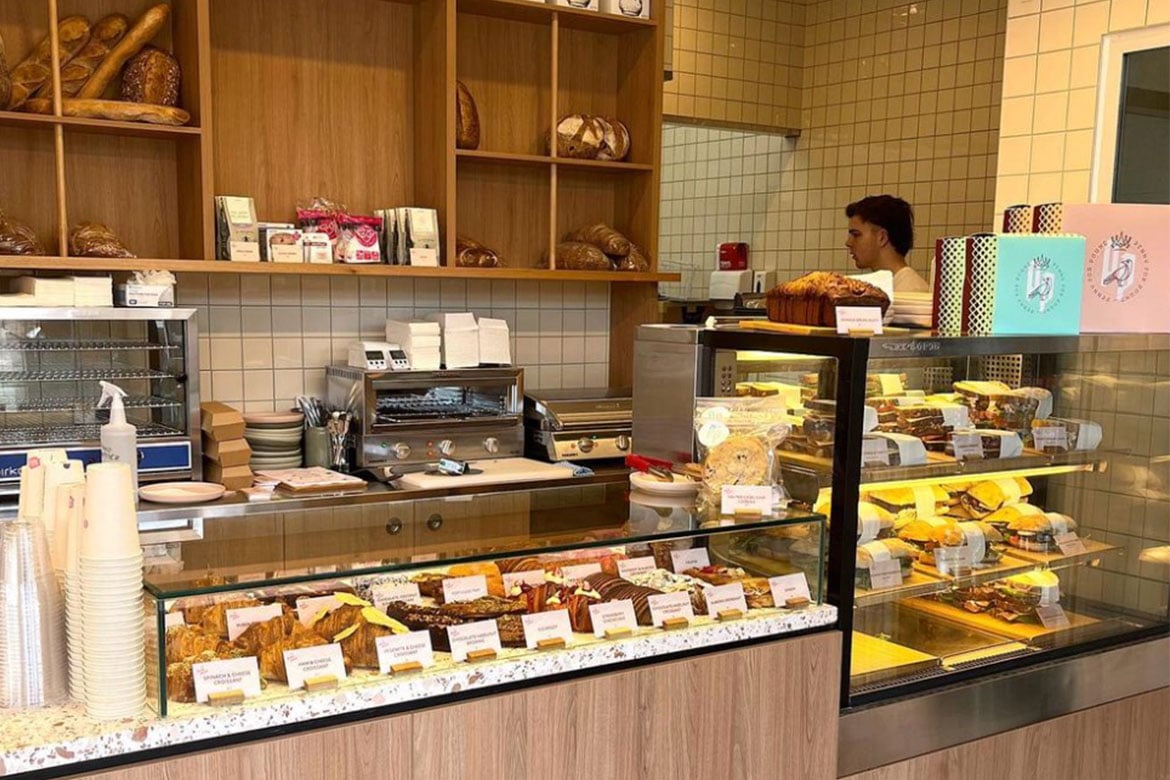 The interior of a bakery/cafe; there are big glass cases full of sweet and savoury pastries. Behind is a shelf stacked with bread and a coffee machine. On the right, behind the counter, there is a man. (Penny for Pound)