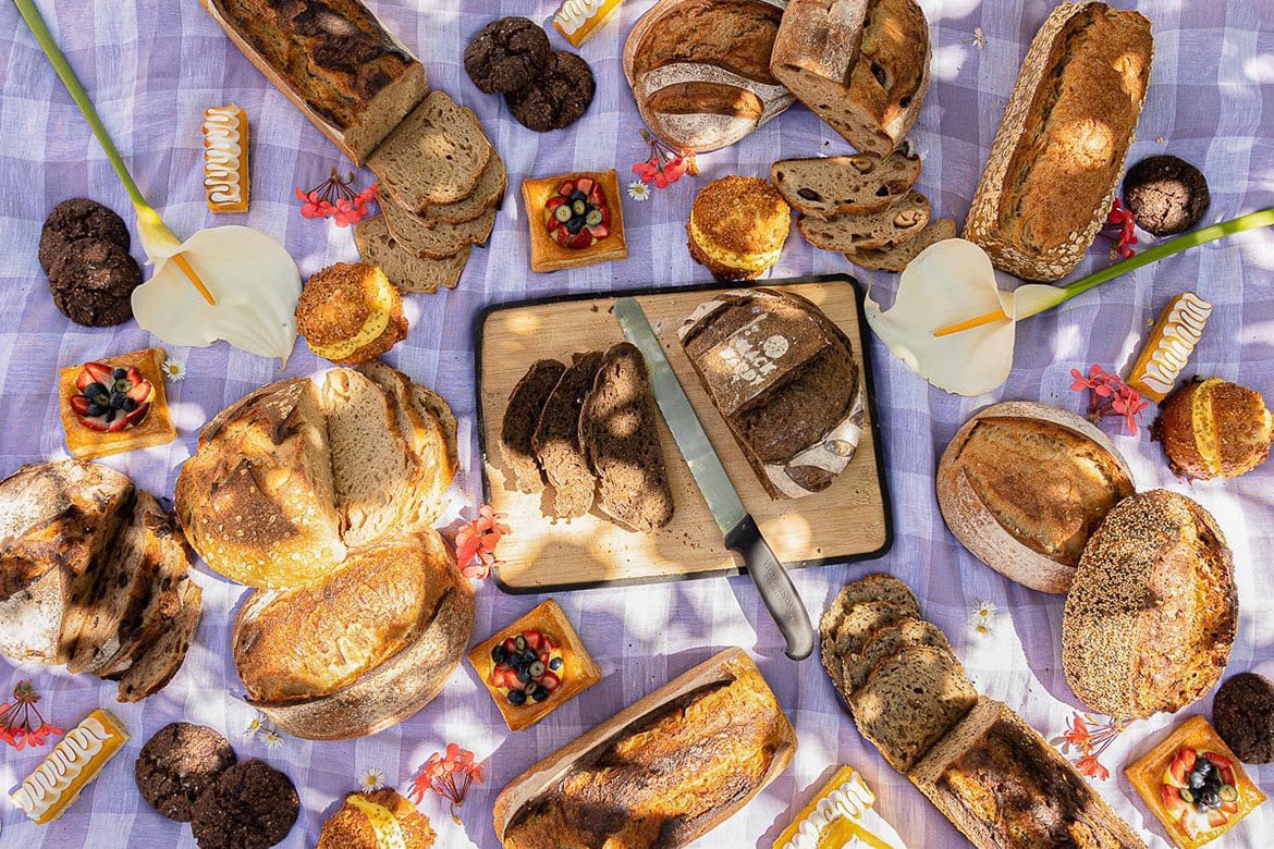 A picnic spread of various loaves of bread, positioned beautifully around a large loaf of bread in the middle of the picnic rug on a chopping board (Back Alley Bakes)