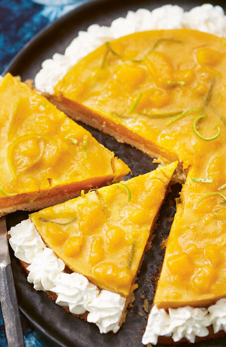 A mango tart sits on a plate resting on a blue crushed velvet tablecloth. Two slices are cut from the tart, resting on the plate.