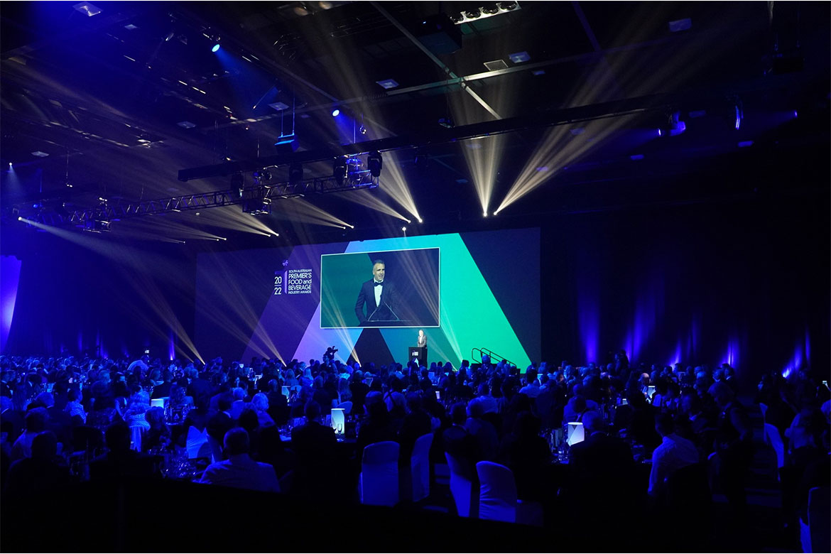 A group of people sit at dinner tables. On the stage is a speaker, a screen behind him shows him in a large image (South Australian Premier's Food and Beverage Industry Awards)
