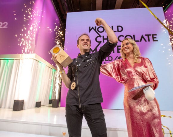 Winner of the World Chocolate Masters, Lluc Crusellas, stands on a stage holding aloft his trophy. A woman in a pink dress stands next to him.