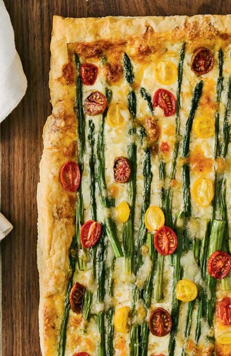 Asparagus tomato and cheese tart sits on a wooden chopping board; cloth napkins rest beside it