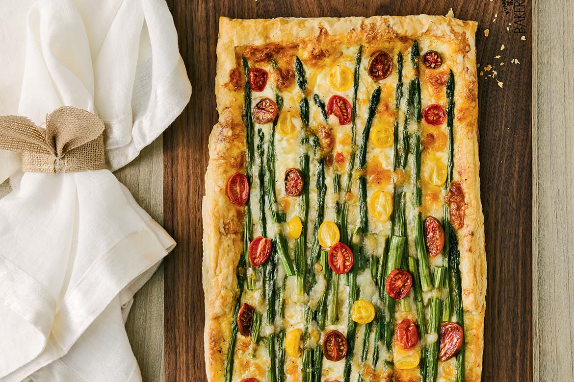 Asparagus tomato and cheese tart sits on a wooden chopping board; cloth napkins rest beside it