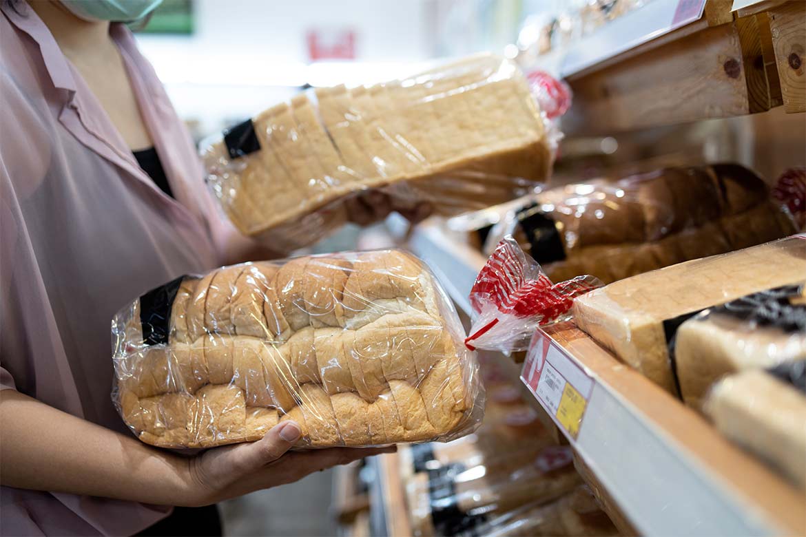 A woman stands at a bread shelf in a supermarket with a loaf of bread in each hand (food waste)