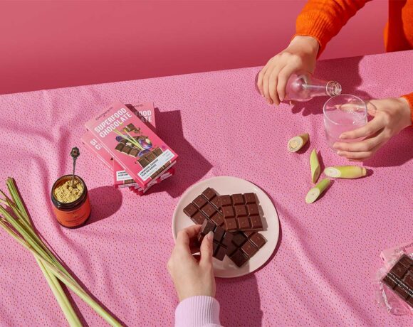 A brightpink. tablecloth; a plate full of chocolate. Someone is pouring a glass of water. THere are stalks of lemongrass. (Loving Earth)