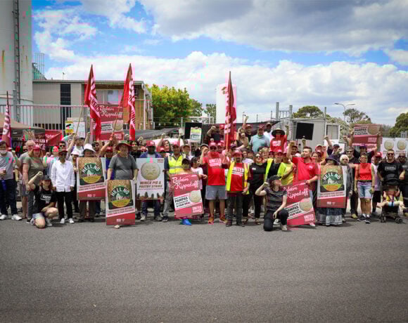 A group of striking Pampas Pastry workers stands in front of a factory holding signs and placards in protest