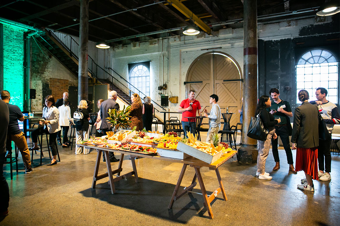 A large display of fresh fruit and veg, breads, etc., sits in the middle of a conference room surrounded by people talking to each other (thinkfood LIVE)