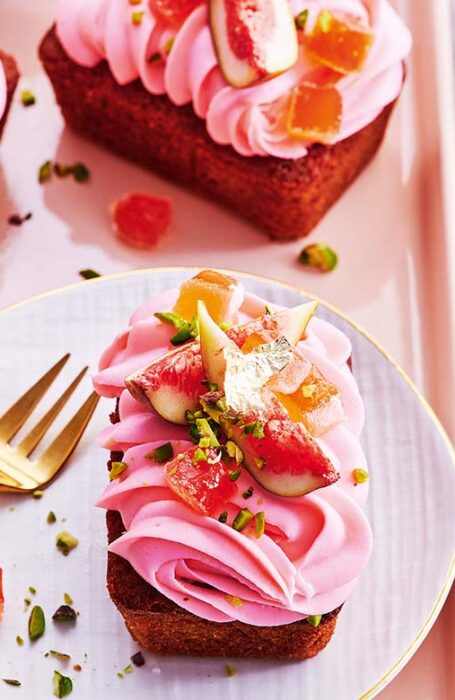 Several rose and fig love cakes on a pink tray, one siting on a plate with a dainty gold cake fork next to it