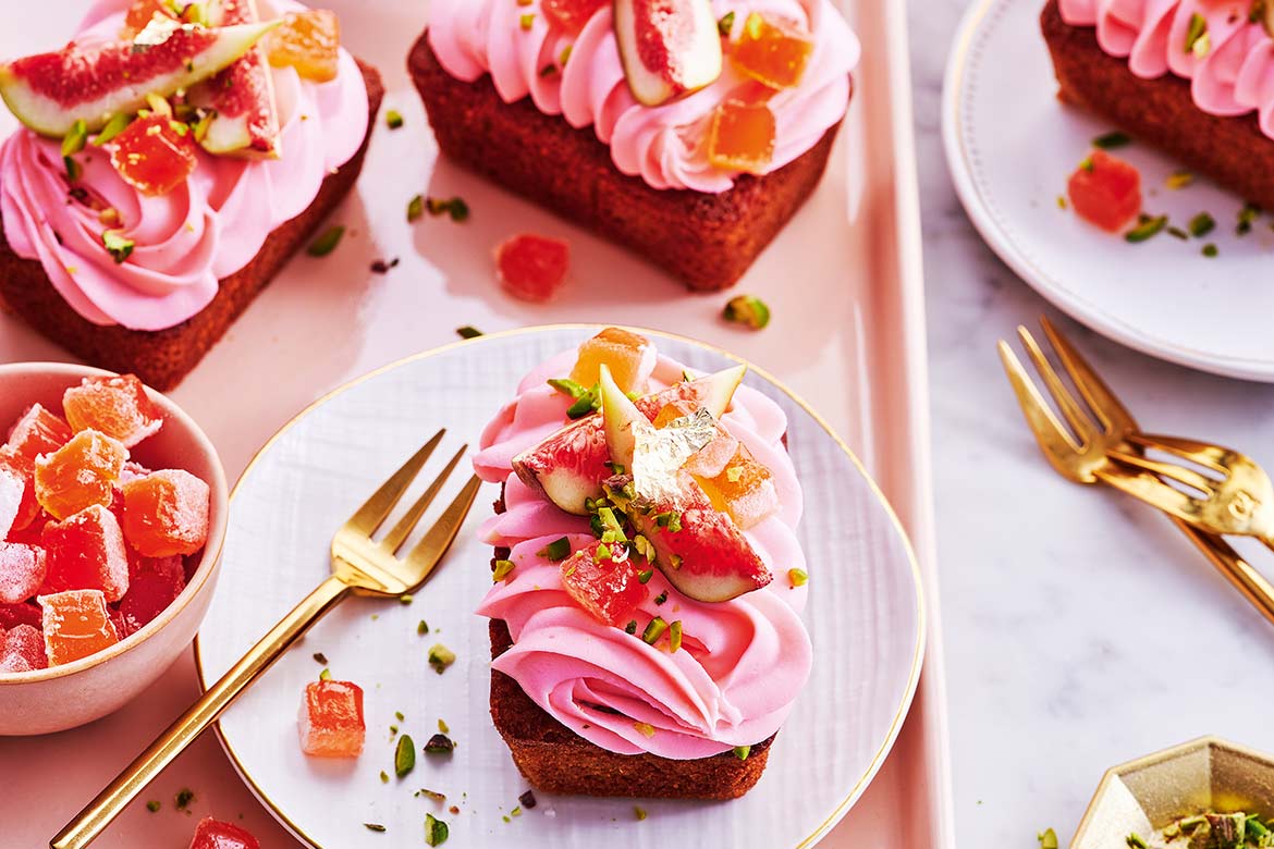 Several rose and fig love cakes on a pink tray, one siting on a plate with a dainty gold cake fork next to it