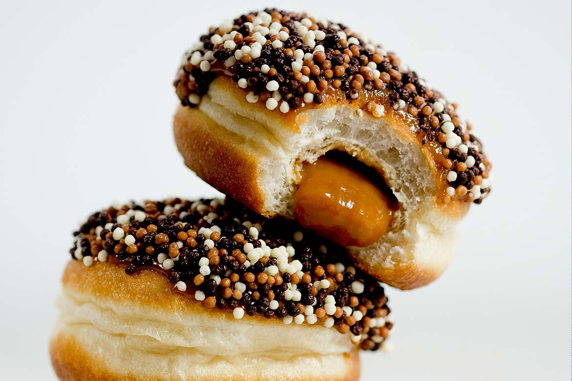 Two delicious-looking stuffed doughnuts stacked on each other. They are topped with chocolate and sprinkles, the top one has a bite taken out of it, showing the oozy, gooey caramel inside (Millionaires Caramel)