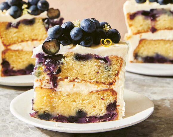 A slice of double-layered sour cream blueberry cake with vanilla mascarpone icing sits on a plate, two slices on plates are in the background