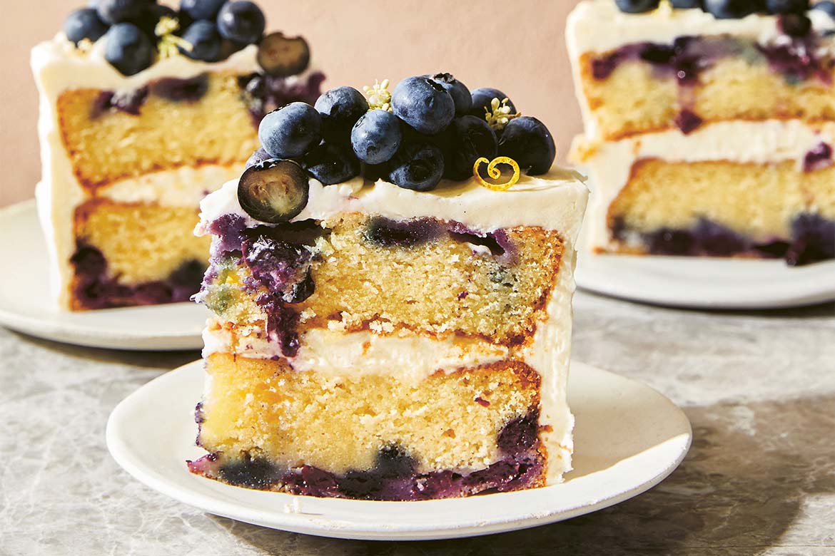 A slice of double-layered sour cream blueberry cake with vanilla mascarpone icing sits on a plate, two slices on plates are in the background