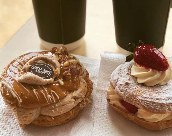 Two filled paris-brests, one with a strawberry on top, one with an Uncle Bob's Bakery disc sit on a table in front of two takeaway coffee cups