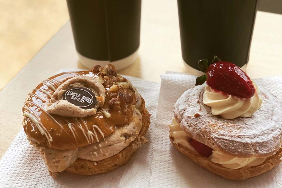 Two filled paris-brests, one with a strawberry on top, one with an Uncle Bob's Bakery disc sit on a table in front of two takeaway coffee cups