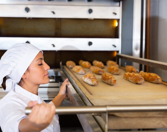 Young Hispanic woman in white uniform working in bakery, pulling freshly baked baguettes out of industrial oven (flexible)