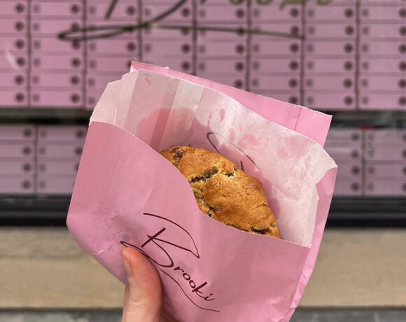 a hand holding a choc chip cookie in a pink Brooki Bakehouse bag in front of the Brooki Bakehouse glass shopfront