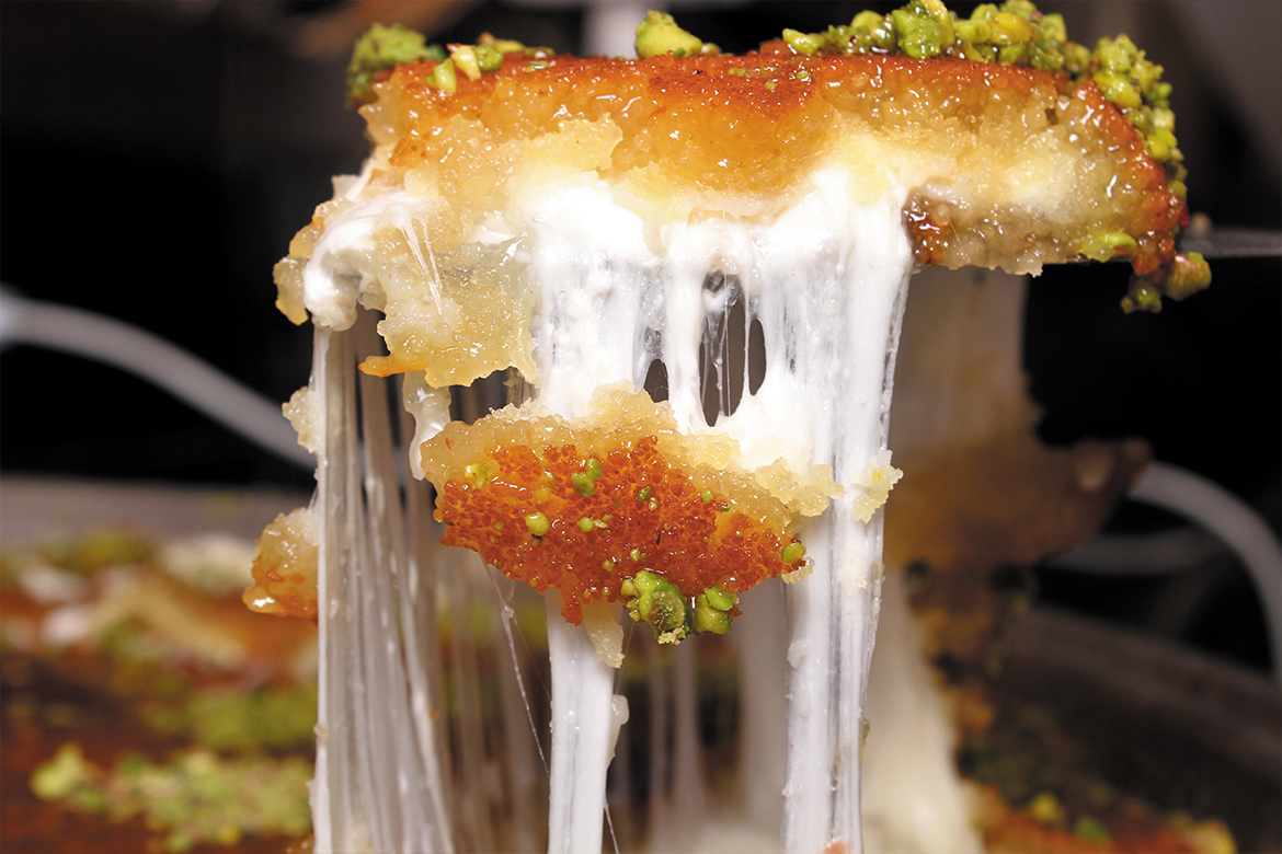 a dish of knafeh being broken, someone is lifting a piece out using a spoon, the cheese is very stretchy (Knafeh Nabulseyeh)