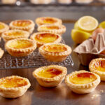 Portuguese custard tarts sitting on a metal bench; some are directly on the bench, some are on a wire cooling rack. To the right is an egg carton with 3 eggs in it, in the background are a couple of cut up lemons