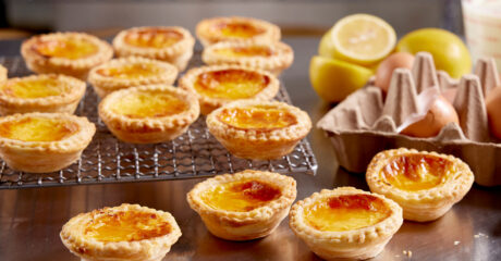 Portuguese custard tarts sitting on a metal bench; some are directly on the bench, some are on a wire cooling rack. To the right is an egg carton with 3 eggs in it, in the background are a couple of cut up lemons