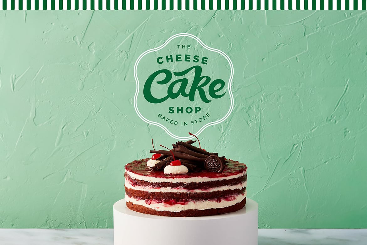 The Cheesecake Shop sign on a pale green background with a black forest cake in front