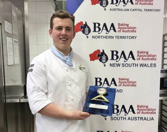 Excellence in Baking NSW winner William Nickl stands in front of a BAA banner holding his trophy. He wears chefs whites and smiles at the camera