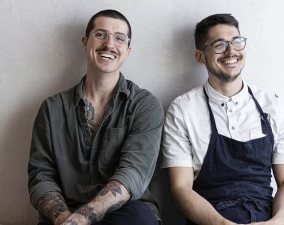 LoDe Pies co-owners Lorenzo Librino and Benji Spencer will be stepping back from the business. Image is two men, one in a grey shirt and the other in a blue apron and white shirt, sitting and leaning on a grey wall.