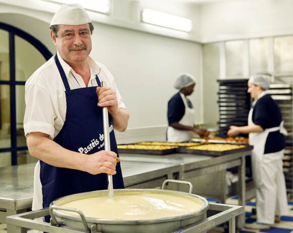 A baker with a grey moustache stirs a large pot of custard. He's looking at the camera.