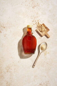 A bottle of maple syrup lies on its side, next to a spoon of maple sugar and a pile of maple flakes.