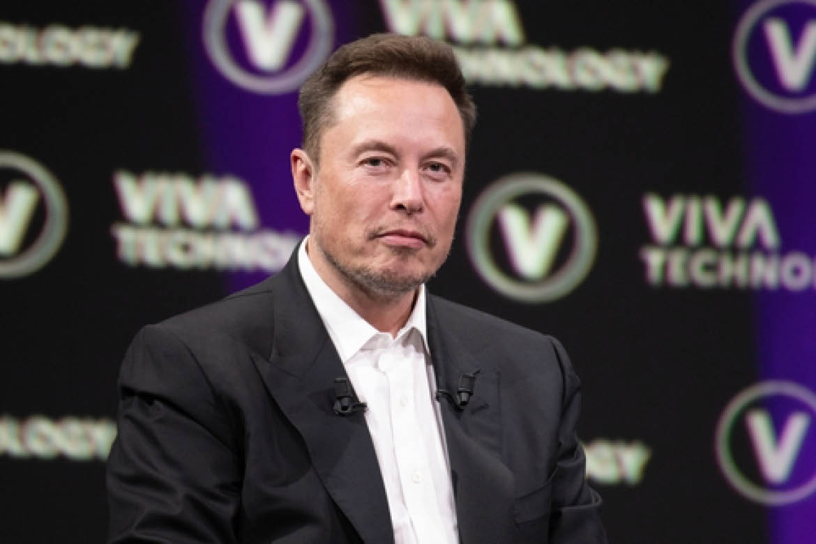 Tesla owner Elon Musk is pictured in a dark suit with a white shirt. He's in front of a dark background.