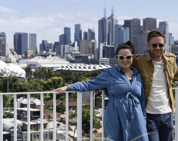 Baker Bleu founders Mike and Mia Russell stand against a white metal fence. It's a sunny day and the Melbourne CBD skyline is behind them. Mia wears a denim dress and sunglasses, Mike wears a white shirt and khaki jacket.. Both are smiling at the camera.