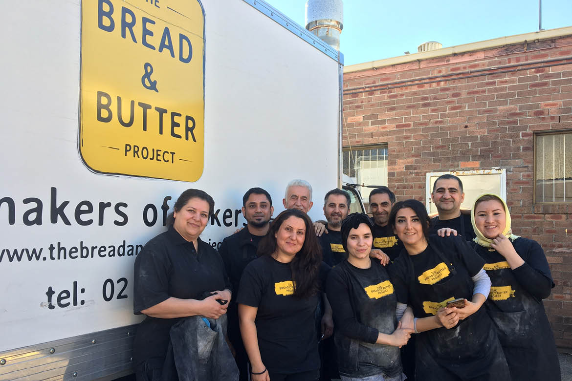 The Bread and Butter Project team poses in front of a white sign that bears the yellow Bread and Butter Project sign. Each team member wears a black shirt.