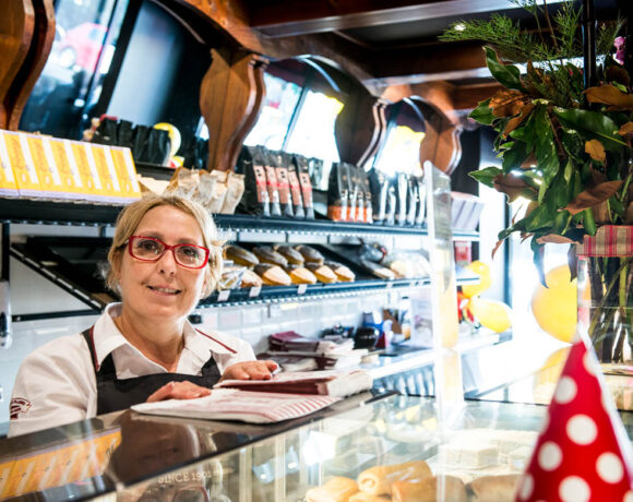 Ferguson Plarre Bakehoue franchise Marie Burrafato stands behind the glass counter. She's wearing red glasses, a white shirt and a dark apron. Her blonde hair is tied up. She's smiling at the camera.