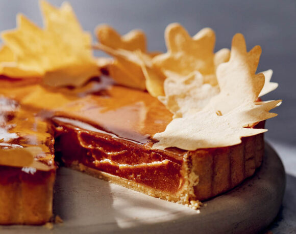 Sweet potato pie sits on a wooden cutting board. A piece has been cut out. Decorative pastry leaves are scattered on top.