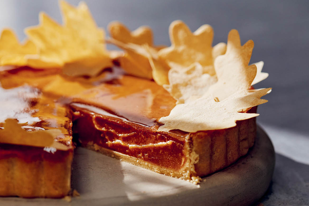 Sweet potato pie sits on a wooden cutting board. A piece has been cut out. Decorative pastry leaves are scattered on top.