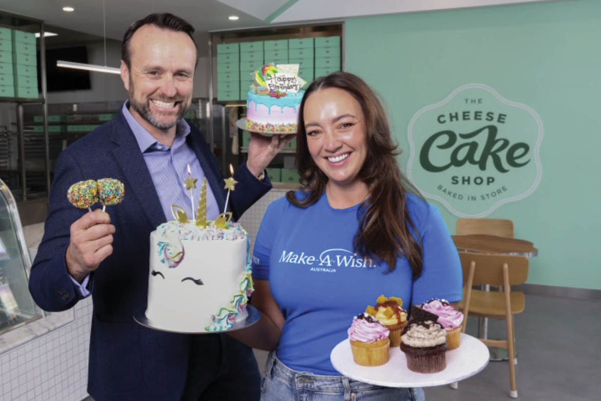 The Cheesecake Shop and Make-A-Wish foundation have joined forces in a three-year partnership. Pictured is a man with dark hair in a dark blue suit holding a cake decorated like a unicorn. Next to him is a woman with long dark hair. She's wearing a Make-A-Wish foundation blue t-shirt and holding a plat of four cupcakes.