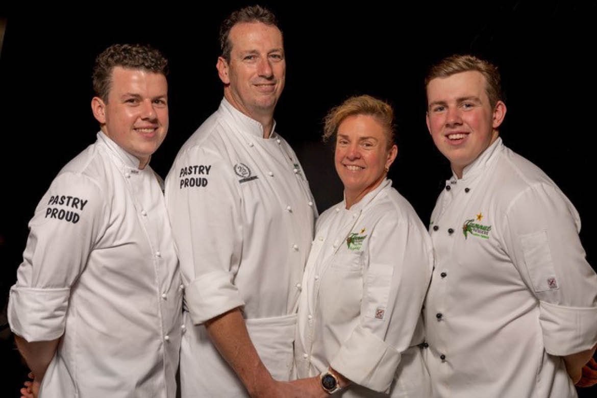 Tracy and Vicki Nickl with their two sons. All four wear chefs whites and are smiling at the camera. There is a black background