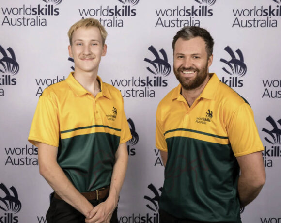 WorldSkills Australia bakery team Lestatt Hammond-Hurst and John Reminis pose in front of a WorldSkills sign. They wear green and gold shirts and smile at the camera.