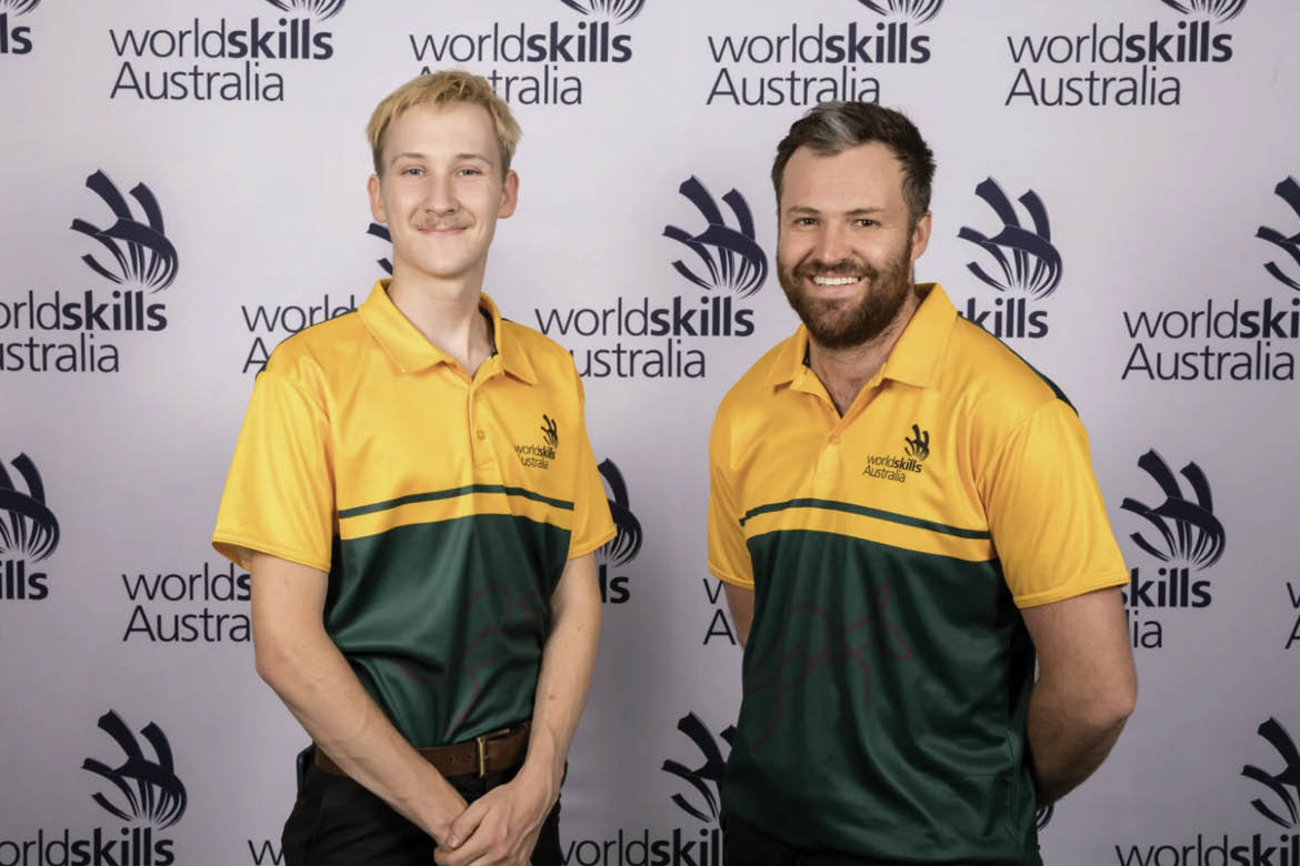 WorldSkills Australia bakery team Lestatt Hammond-Hurst and John Reminis pose in front of a WorldSkills sign. They wear green and gold shirts and smile at the camera.