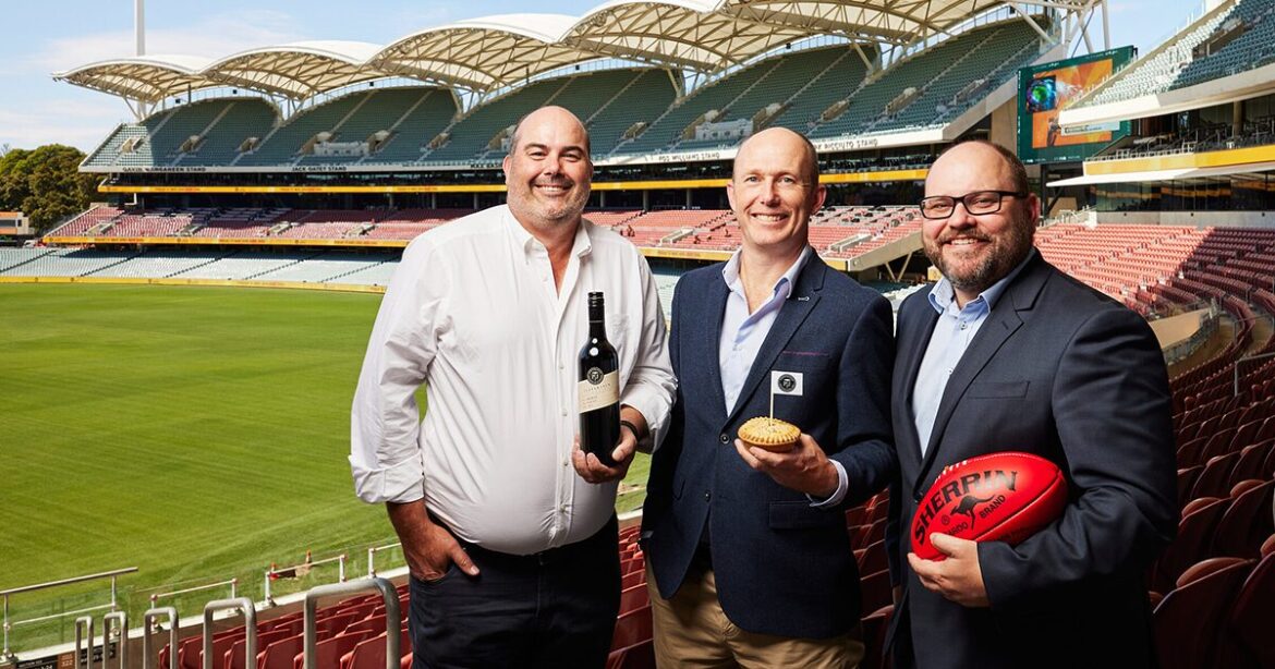 Three business owners of balfours, pepperjacks and AFL standing in Adelaide oval stands holding wine, a footy and a pie