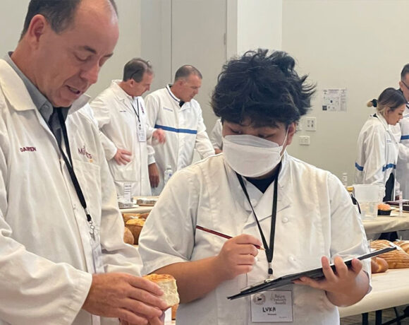 BIEWA competition judges in white coats examine baked good set out in a room full of long tables covered with white tablecloths