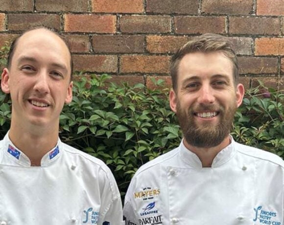 Damien Wright (left) travelled to Italy to compete at the Juniores Pastry World Cup. Pictured is Damien. He has long brown hair tied back in a ponytail. He's wearing a white shirt with branding on it. Next to him is his mentor.