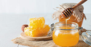 Honeycomb sits on a plate while a jar of honey has a wooden spoon dipped into it.